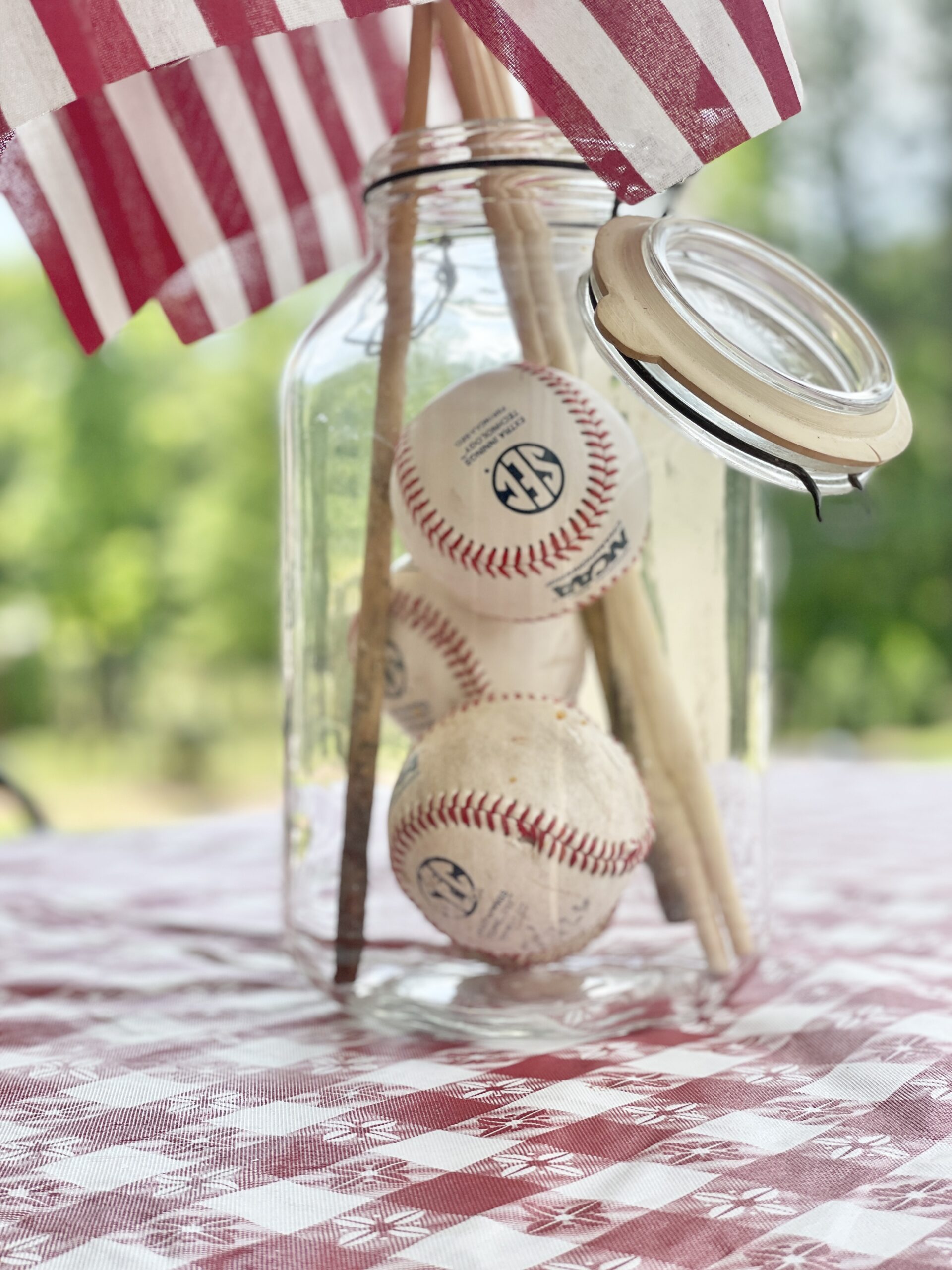 Clear jar filled with baseballs. American flags on sticks tucked into the glass jar with the baseballs. Created a red, white and blue easy centerpiece for the middle of any Summer table. Placed it all on top of a white and red check table cloth.